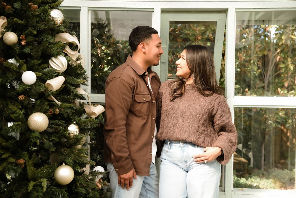 A couple in casual winter outfits shares a romantic moment against a backdrop of white ornaments and greenery.