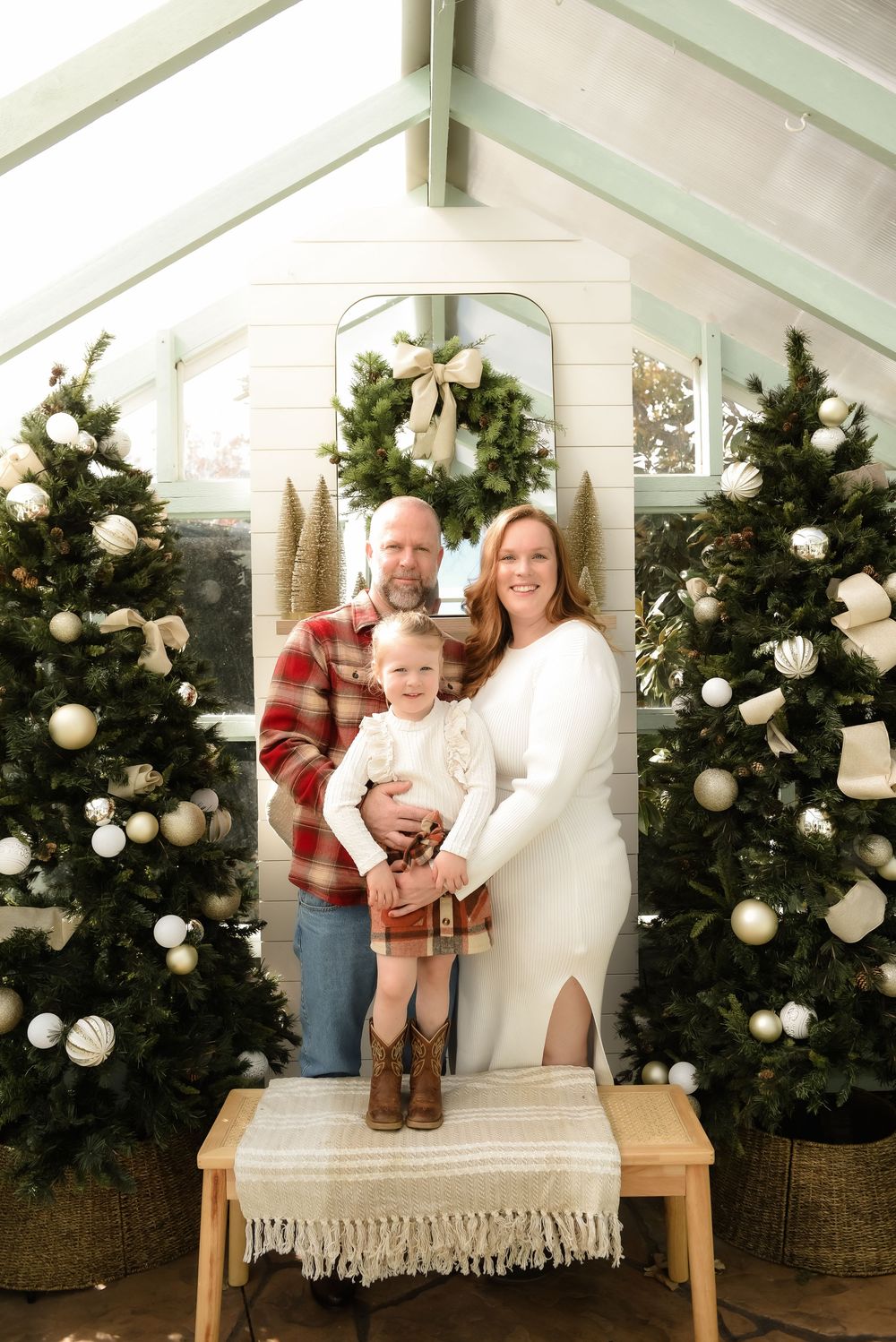 A family poses together on a white bench with decorated Christmas trees in the background for holiday photos.