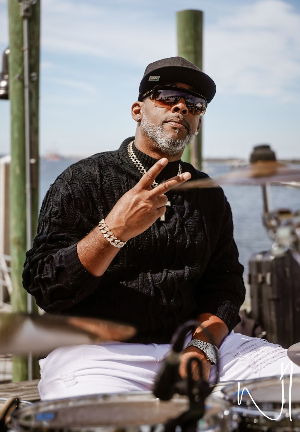 A person in black clothing and sunglasses making a peace sign gesture while seated outdoors.