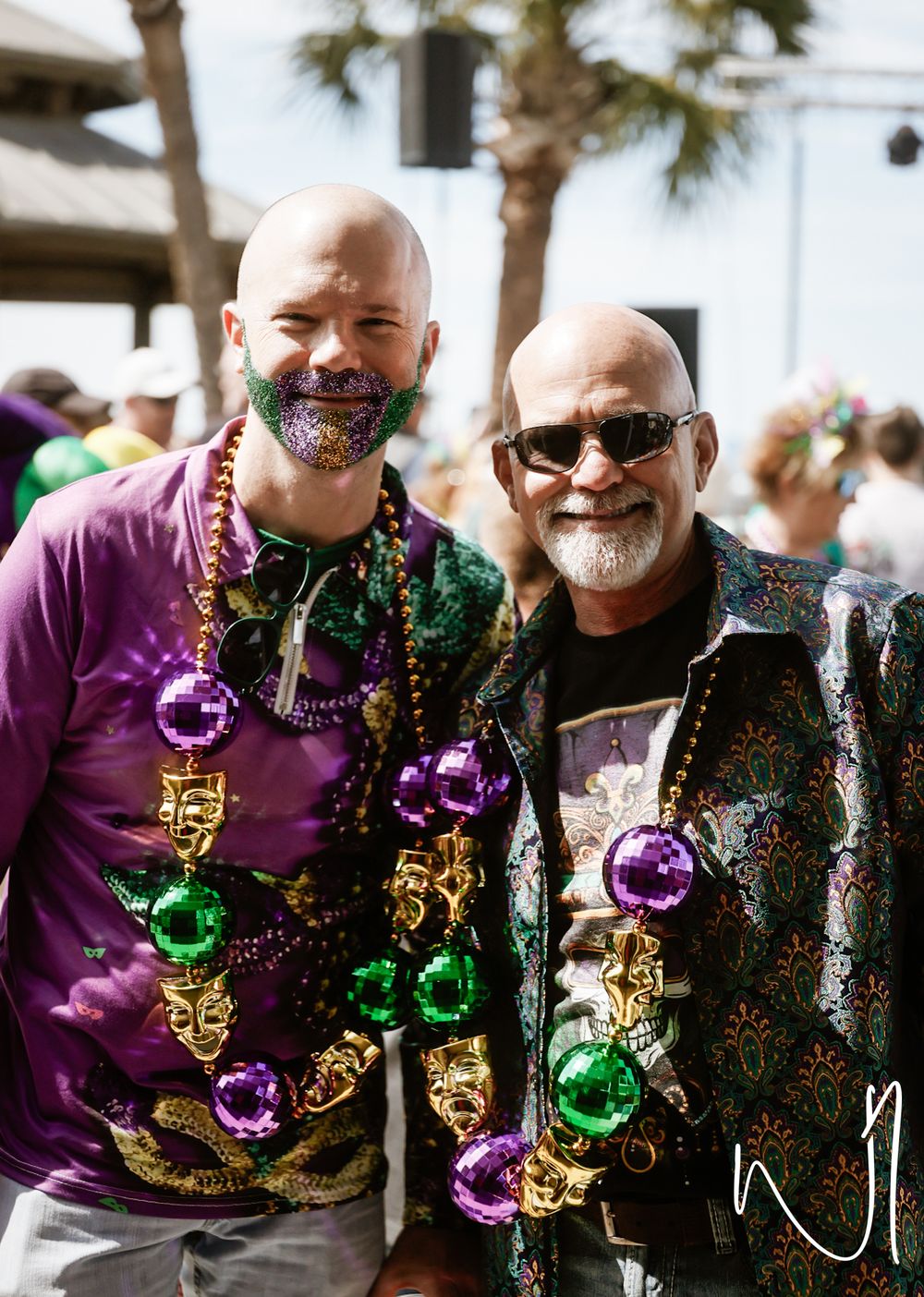 Two people wearing Mardi Gras beads and festive clothing smile together at an outdoor celebration with palm trees.