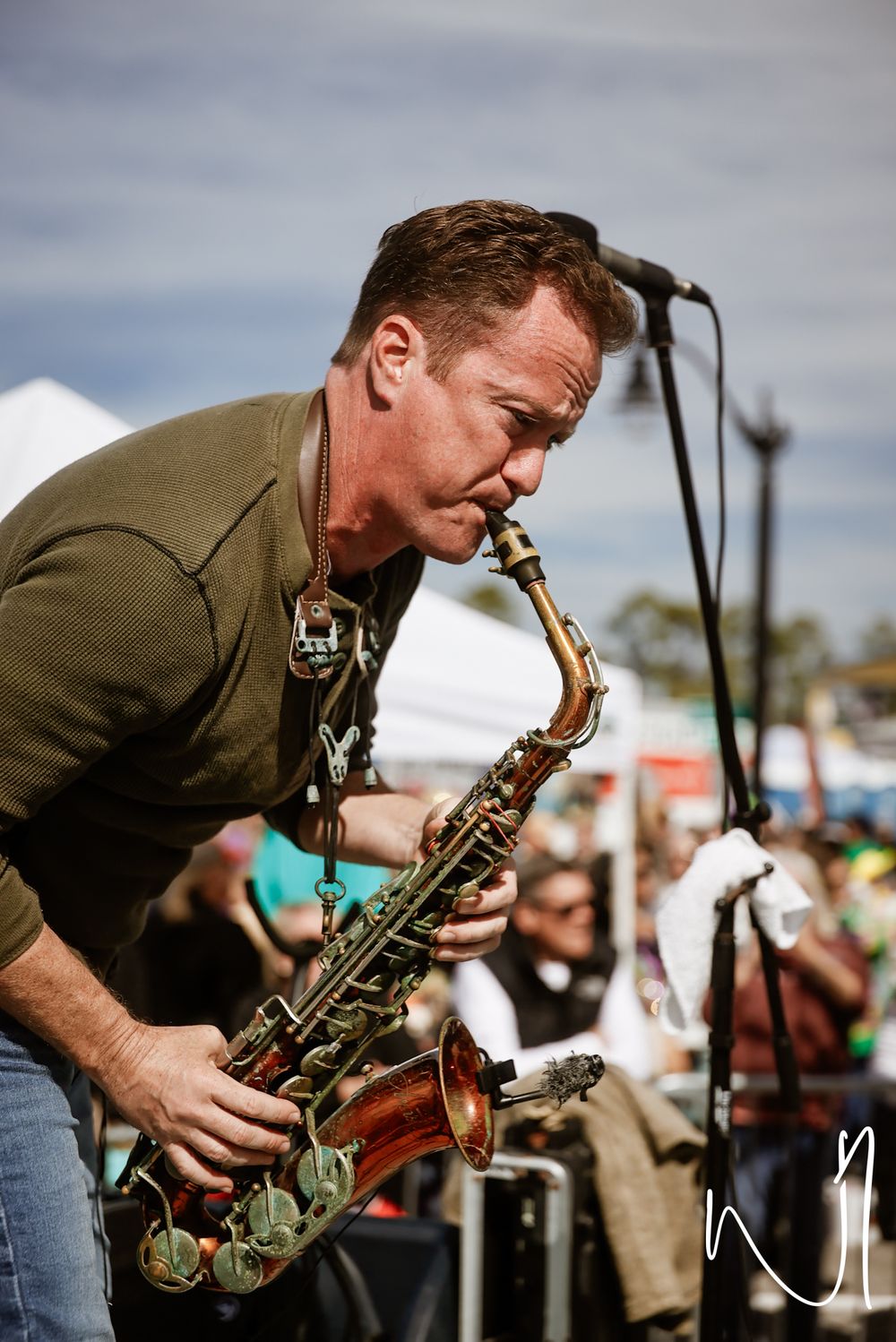 Musician plays saxophone passionately on outdoor stage with microphone and crowd in background.