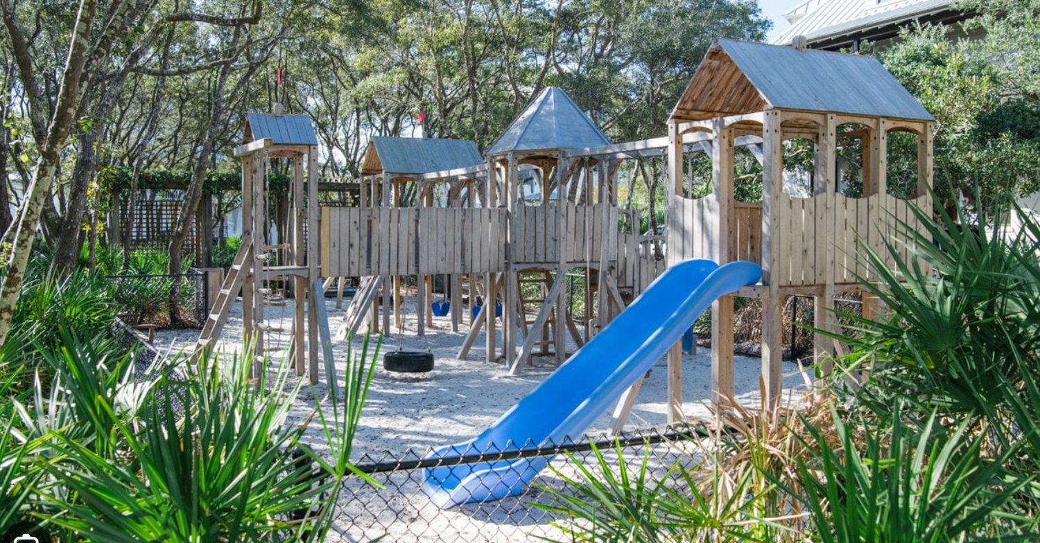 Outdoor playground area with wooden structures, blue slides, and shaded gazebos surrounded by lush greenery.