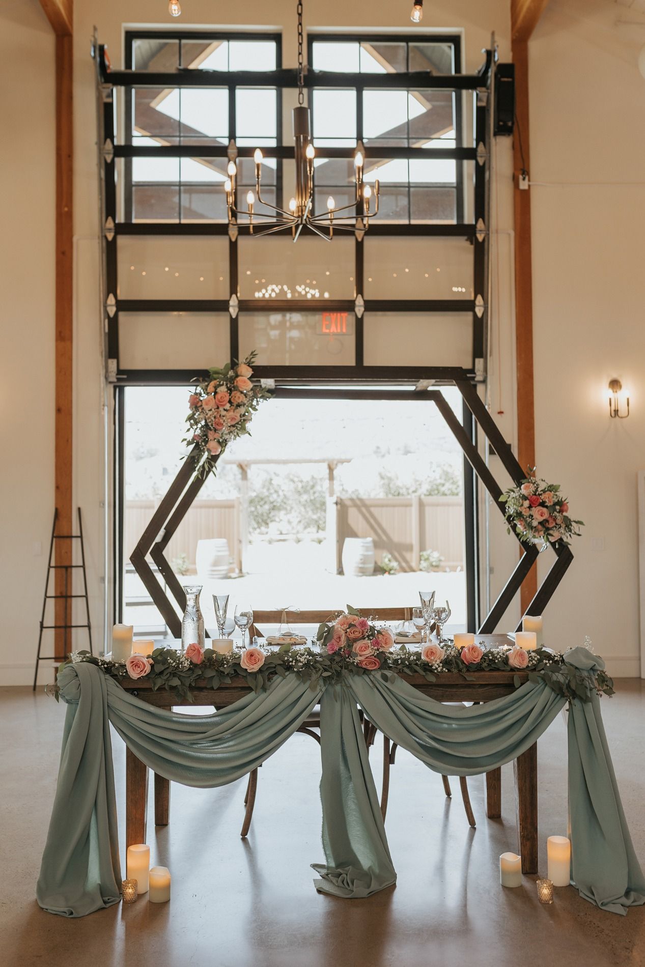 Modern wedding reception setup with hexagonal arch, sage green draping, and floral arrangements in an industrial venue space.