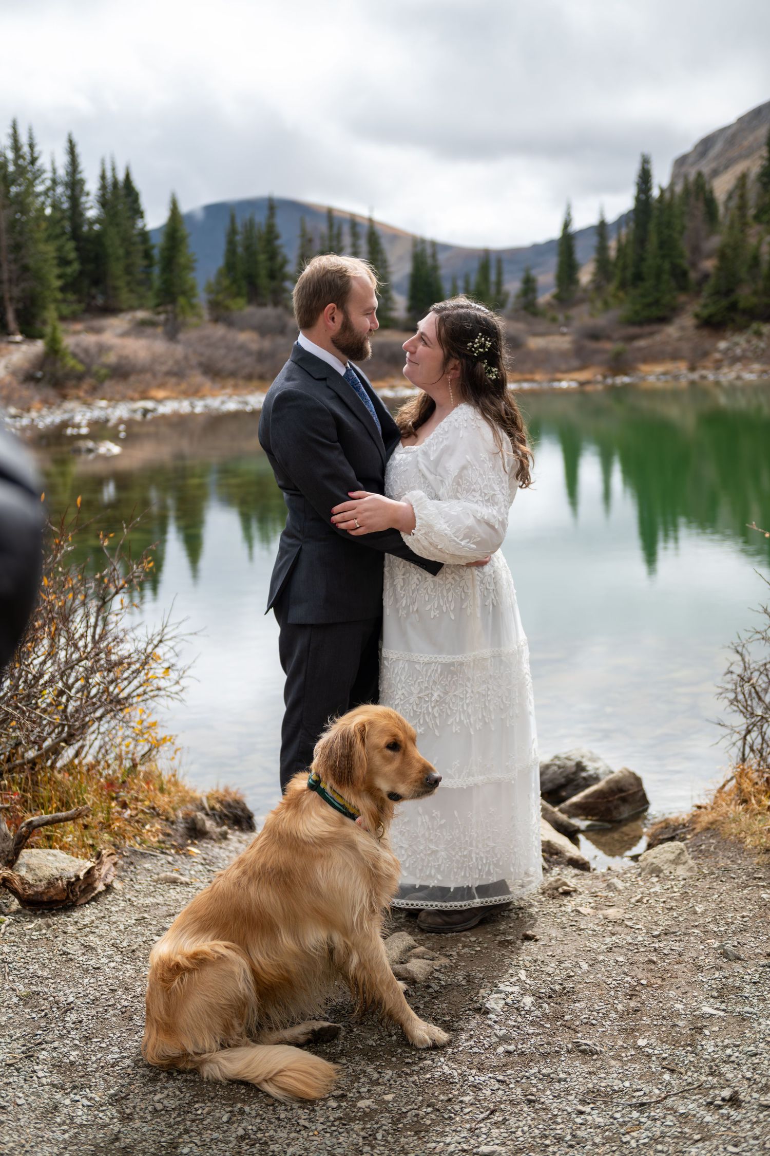 A bride and groom stand chest to chest in an embrace with their dog at their feet. They stand by an alpine lake near Breckenridge.
