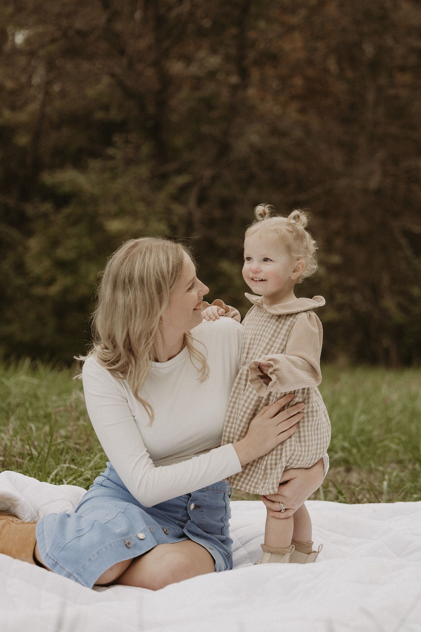 Blonde mother in white top and denim skirt sitting on blanket with smiling toddler in plaid dress.
