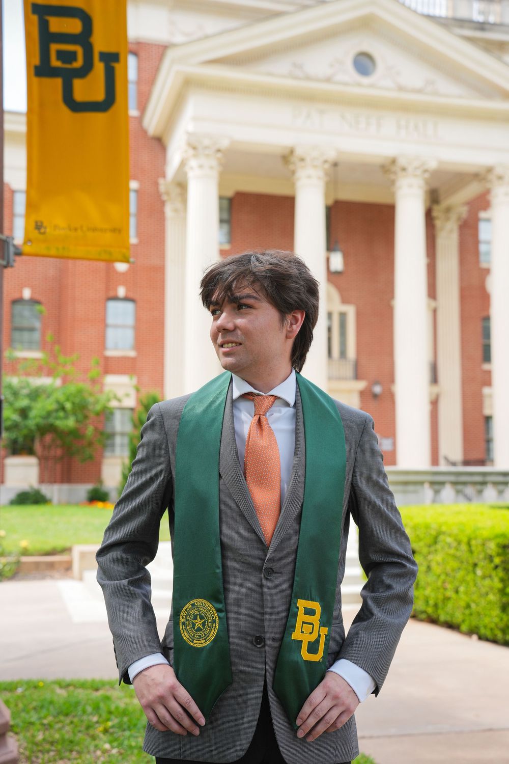 Baylor University graduate in gray suit with green BU stole poses proudly in front of Pat Neff Hall on campus.