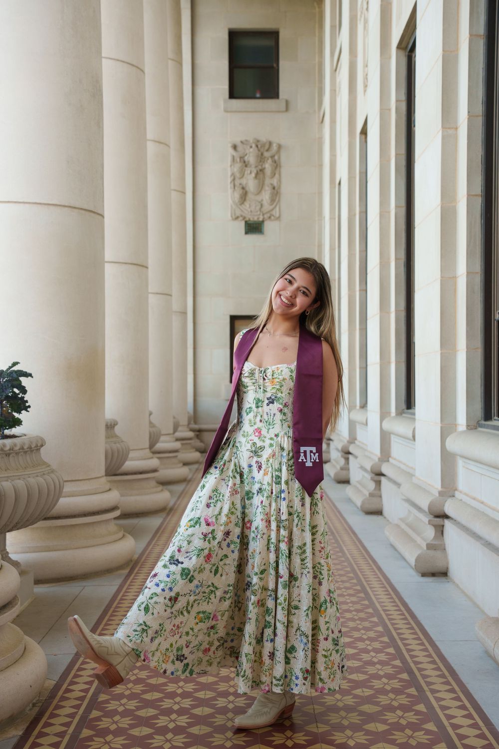 A young woman in a floral dress and red jacket stands in a hallway with columns and decorative architectural details.