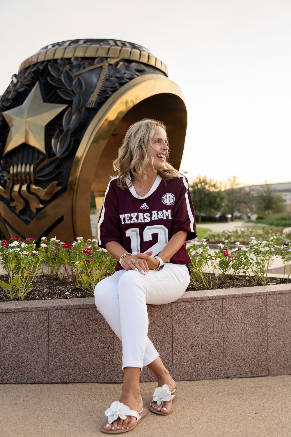 Texas A&M graduate in a maroon jersey sitting on the stone wall at the Aggie Ring Plaza at sunset.