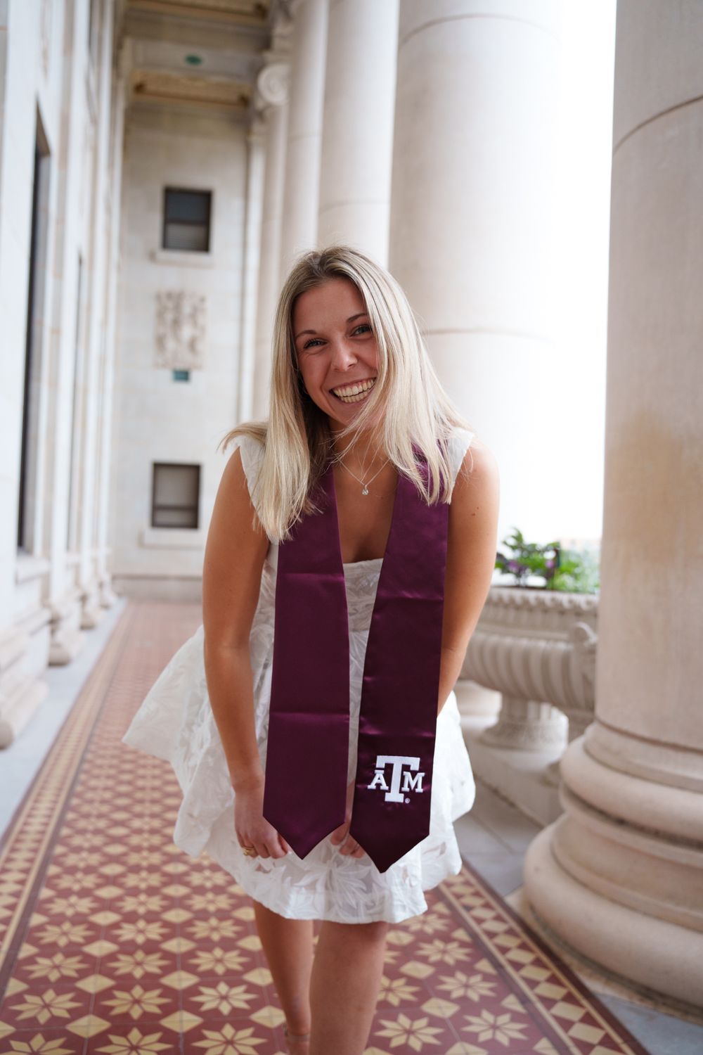 Texas A&M graduate wearing a maroon stole posing along the tiled walkway of the Jack K. Williams Building in College Station.
