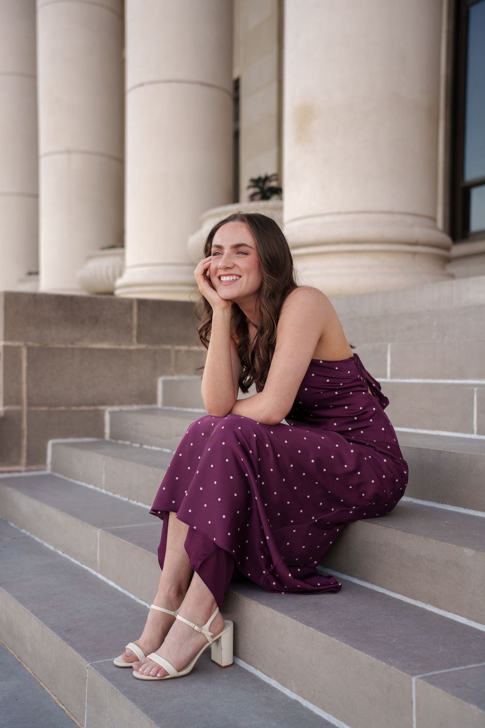 A smiling young woman wearing a purple dress sits on the steps of a building with columns.