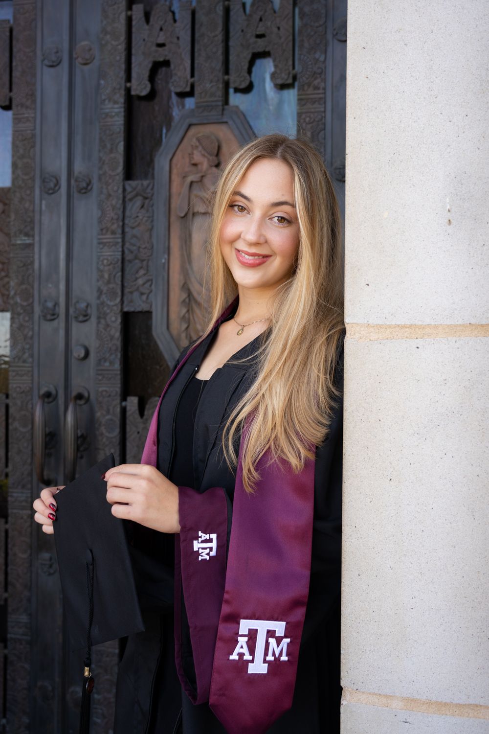 Texas A&M graduate in a maroon sweatshirt posing in front of the dark wooden doors at the Jack K. Williams Building in College Station.
