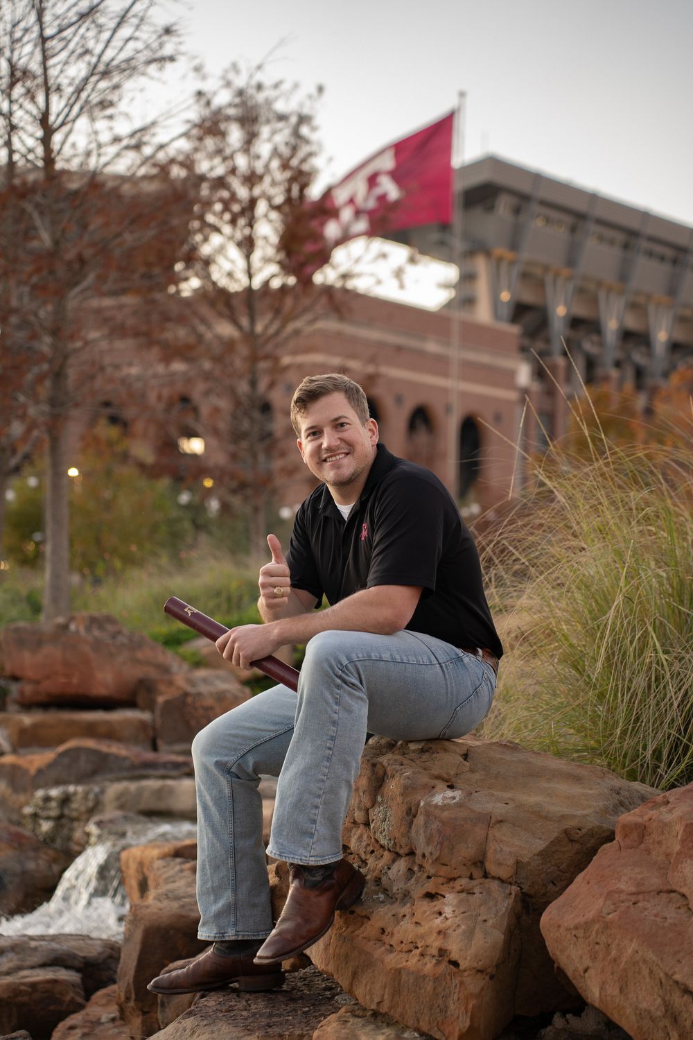 Texas A&M graduate in casual attire sitting on the red rocks near the waterfall at Aggie Park with an Aggie flag in the background.