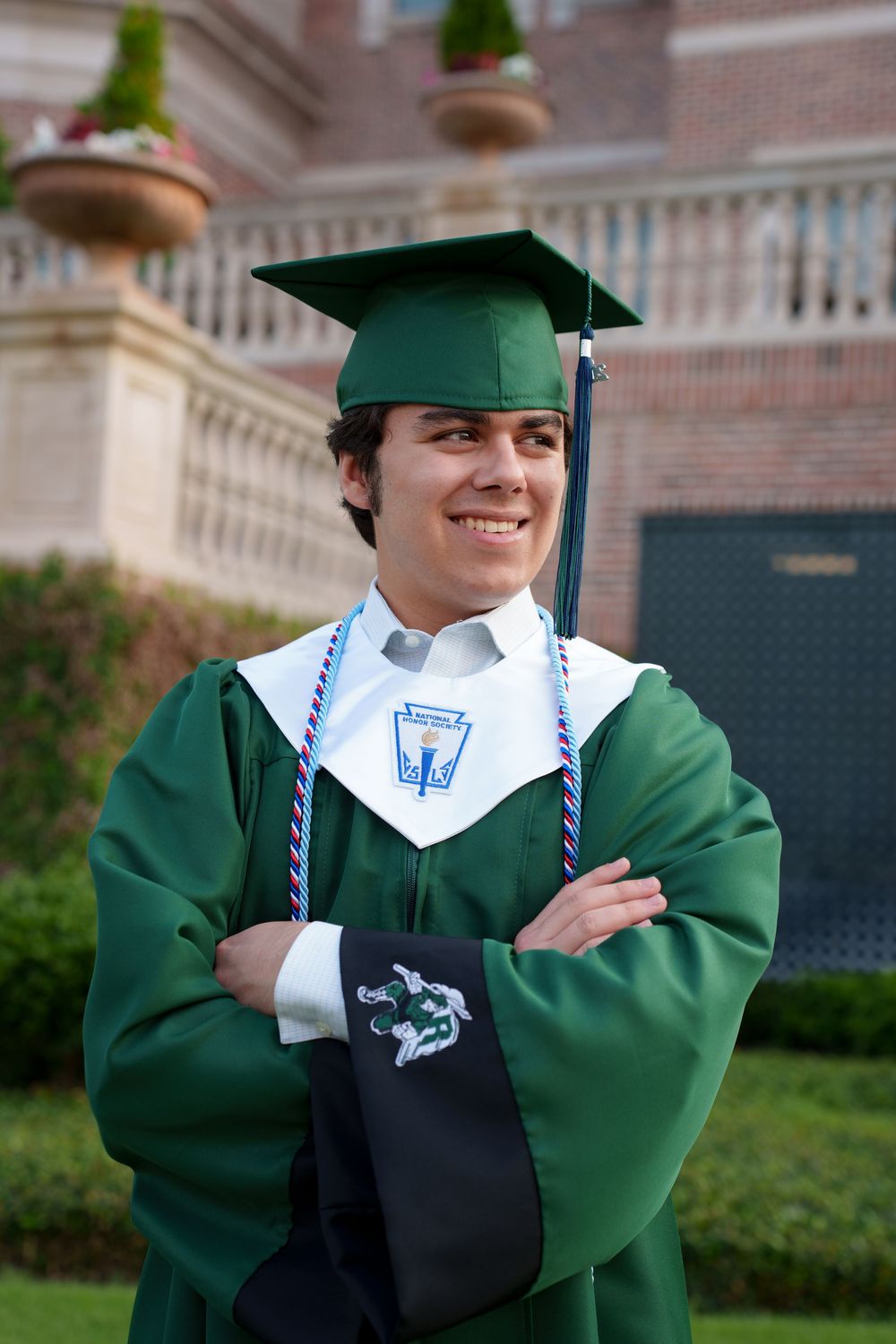 Smiling graduate in green cap and gown with honor cords poses confidently with arms crossed outdoors.