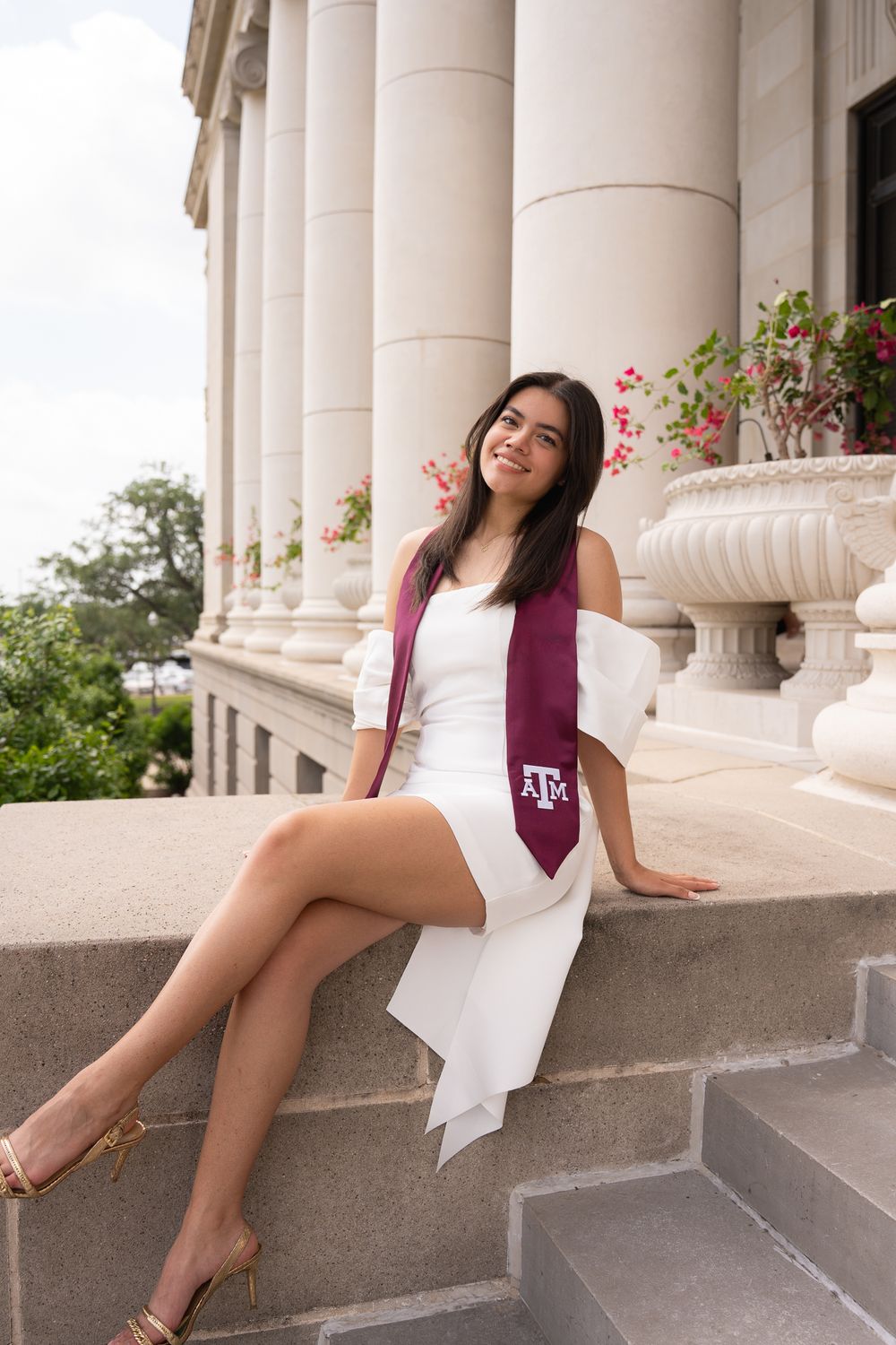 Texas A&M graduate in a white dress and maroon stole sitting on the stone steps of the Jack K. Williams building with flowers.