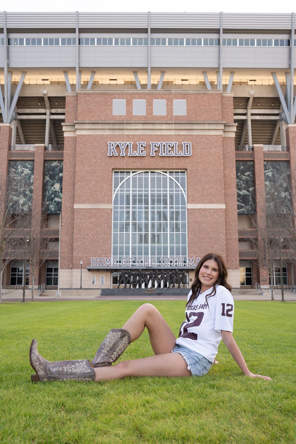 Alt Text: Texas A&M student wearing a jersey and cowboy boots posing on the grass in front of Kyle Field in College Station.