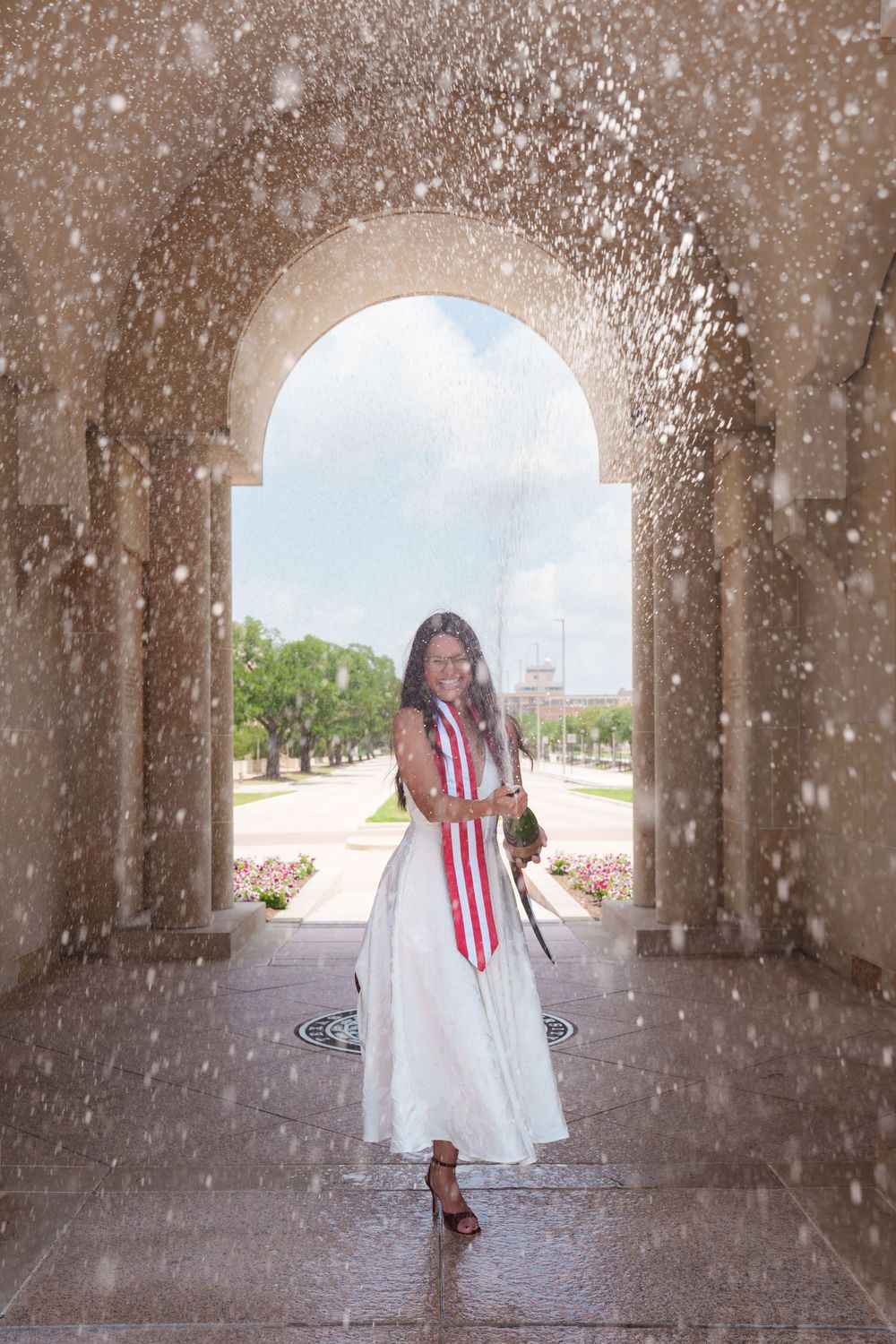 Woman in white dress and red sash stands under stone arch amid water spray, smiling outdoors.