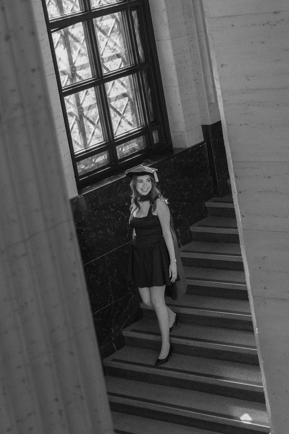 Black and white photo of a young woman in a dark dress posing on elegant marble stairs near a large window.