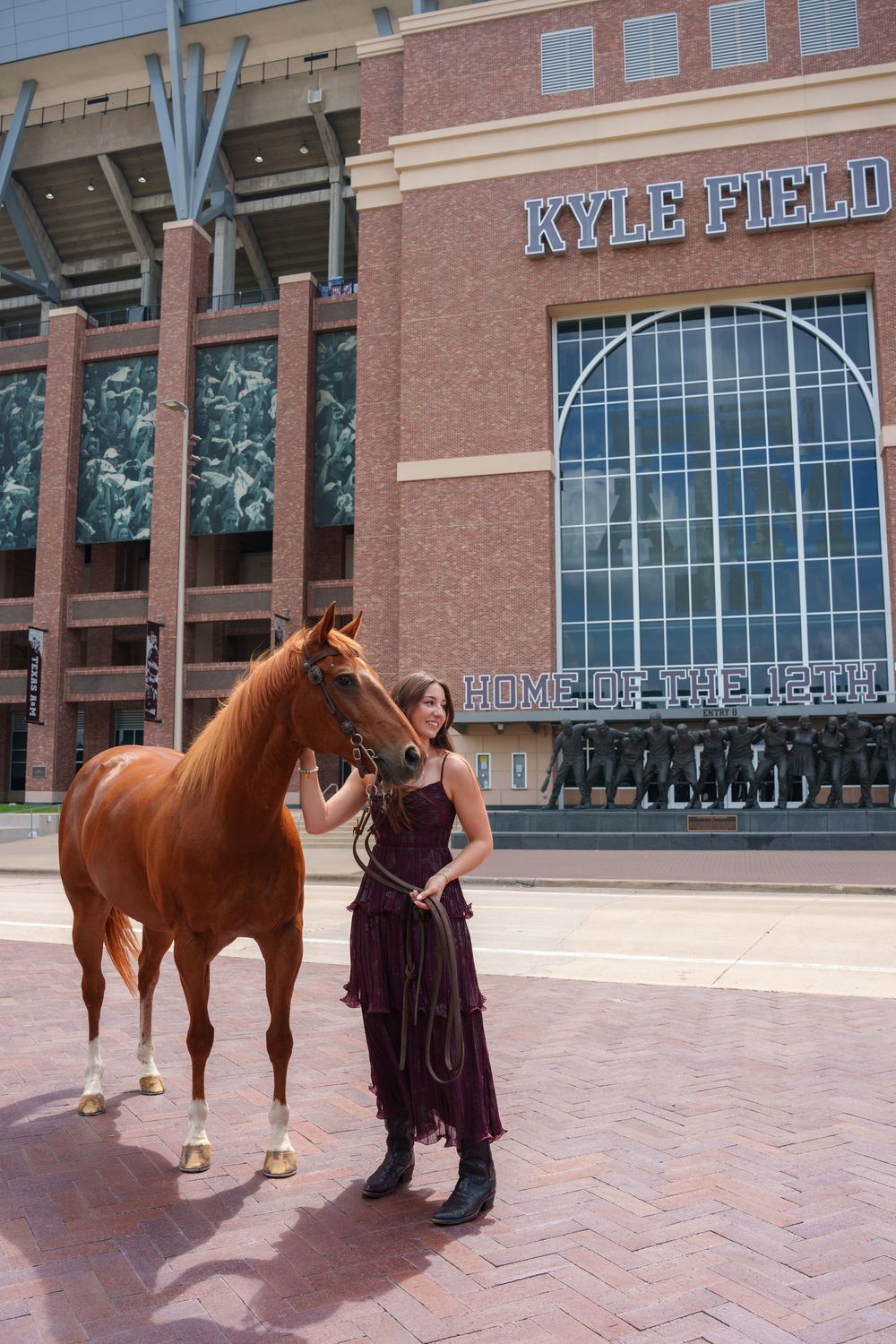 Woman in burgundy dress stands with chestnut horse outside Kyle Field stadium at Texas A&M University.