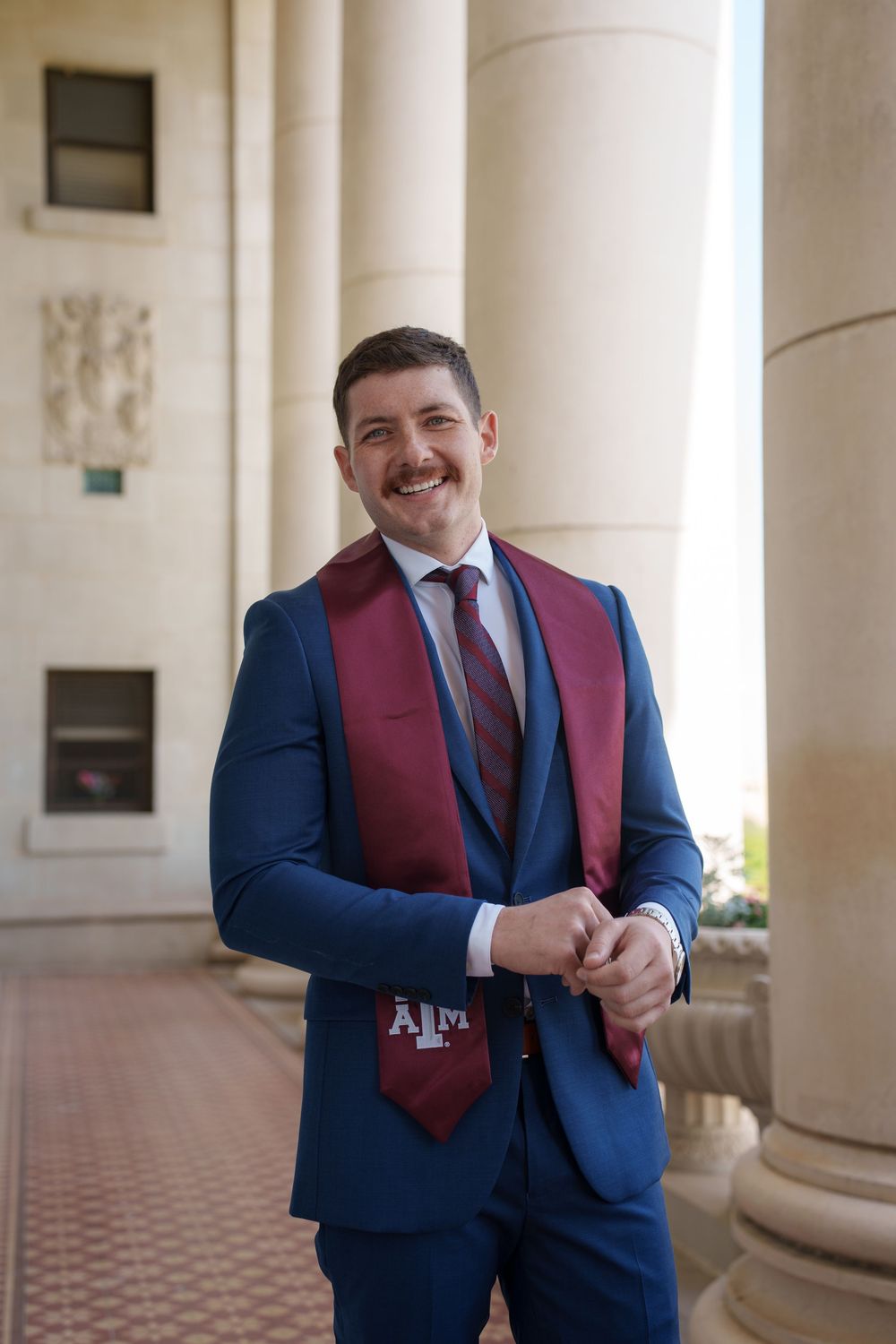 Smiling man in blue suit wearing Texas A&M maroon graduation stole, posing between columns at university.