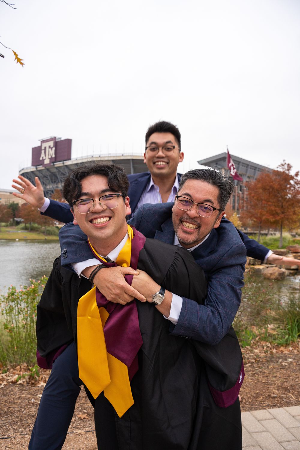 Texas A&M graduate in academic regalia posing playfully with two family members outside a university building in College Station.