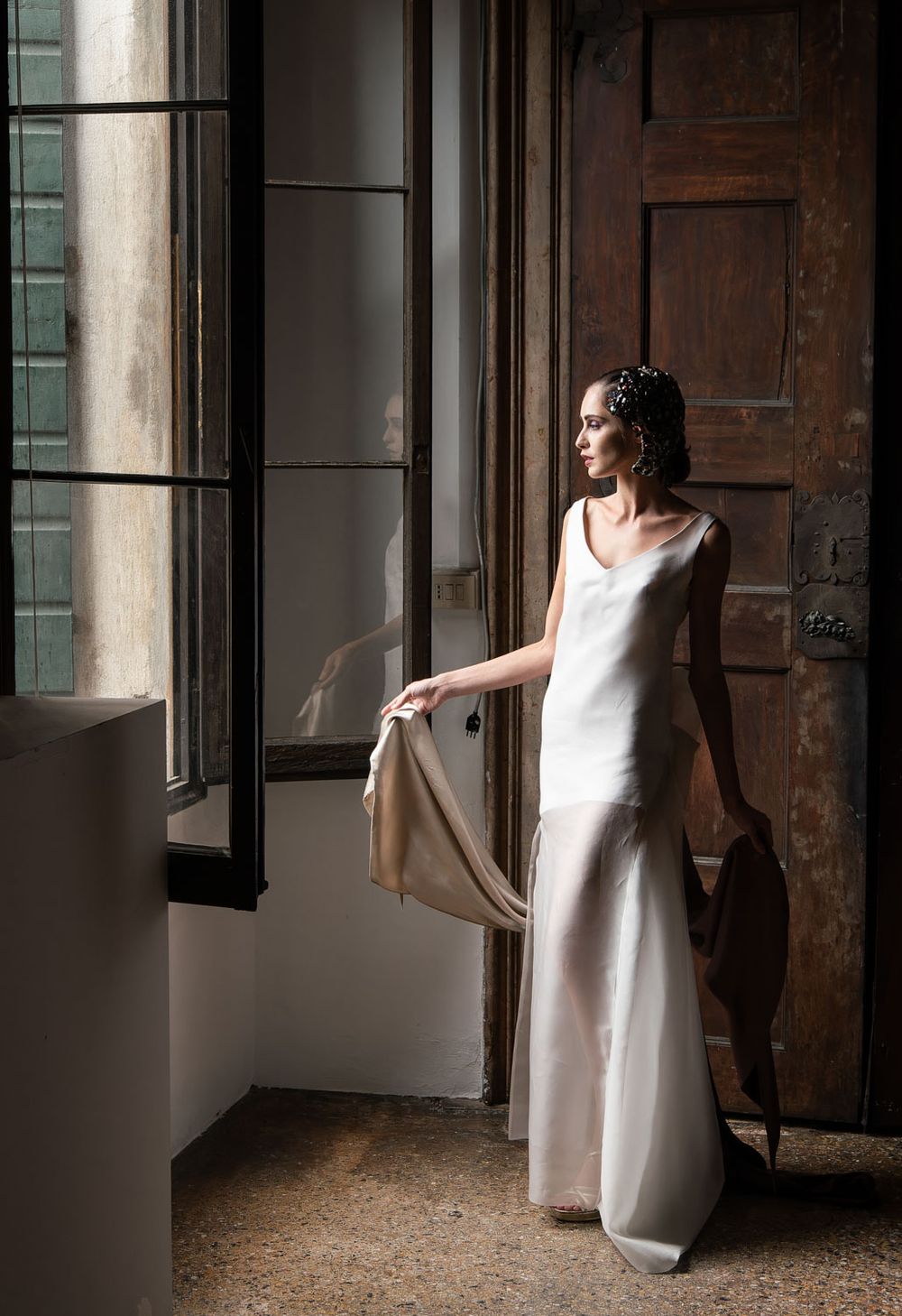 Bride in Venetian palace looks out of window while holding her exquisite dress