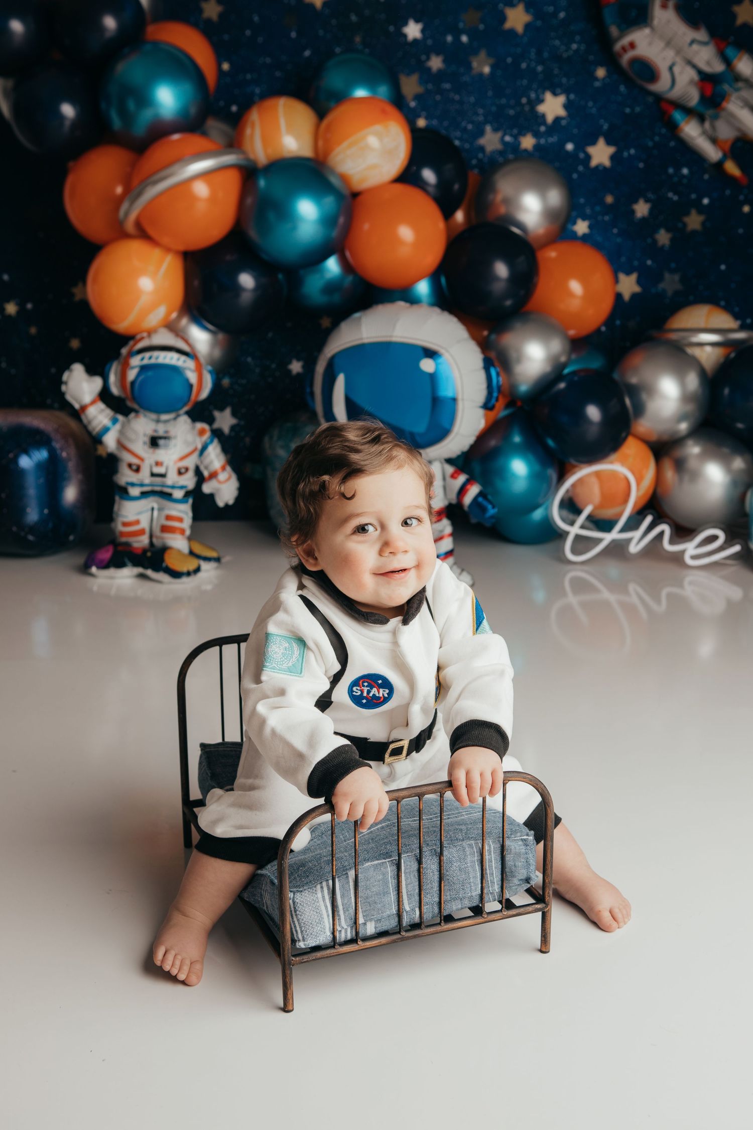 Space-themed baby photo session with astronaut decorations and blue and orange balloon arch.