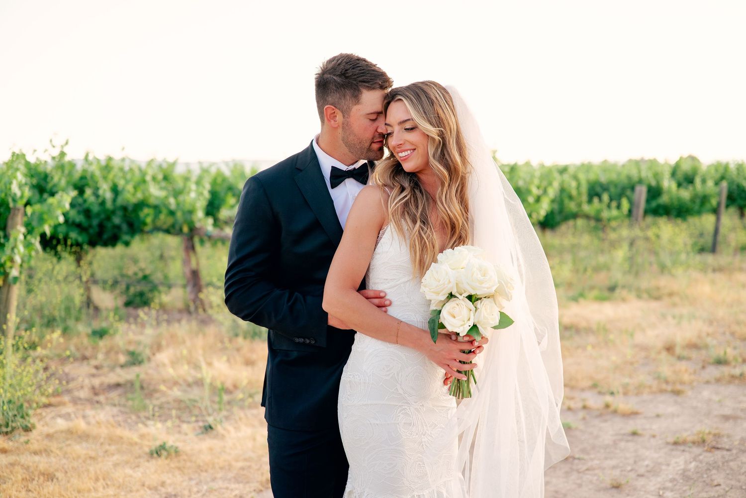 A romantic wedding couple embraces in a scenic vineyard setting during golden hour.