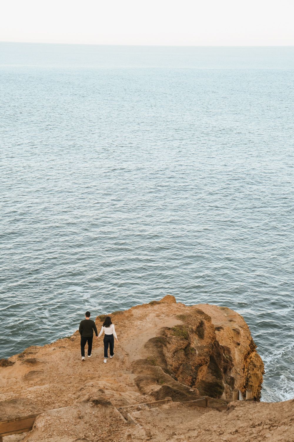 Couple holding hands standing on rocky coastal cliff overlooking calm blue ocean waters.