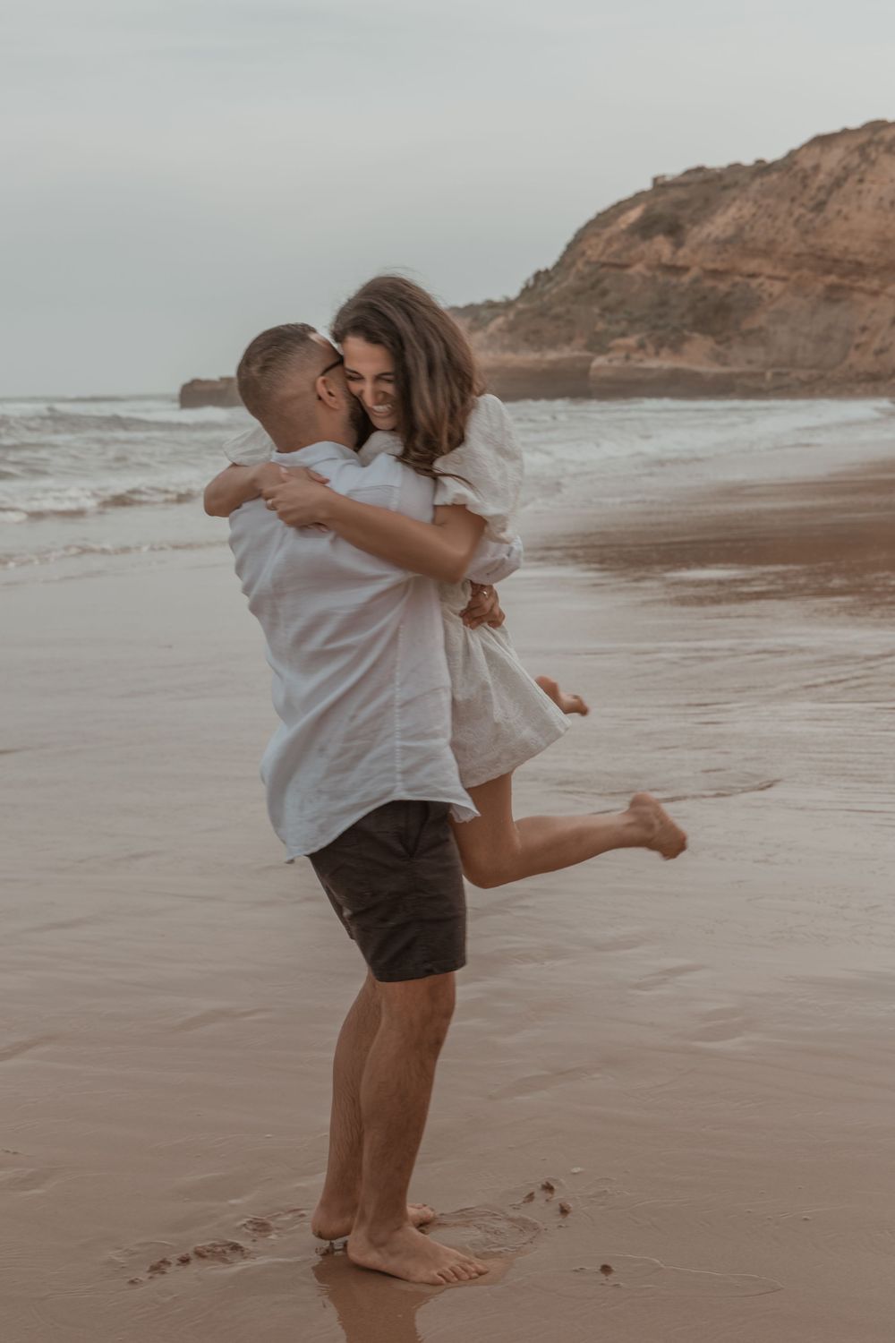 Romantic couple embracing on sandy beach with ocean waves and cliffs in background.