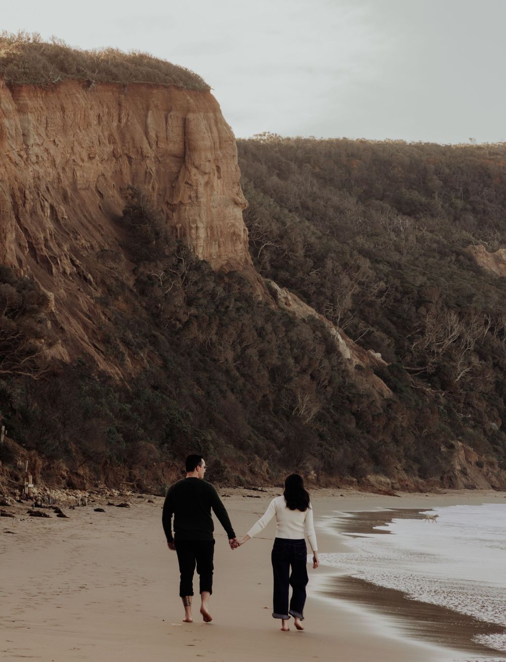Couple holding hands walking barefoot on sandy beach with dramatic sandstone cliffs and lush greenery backdrop.