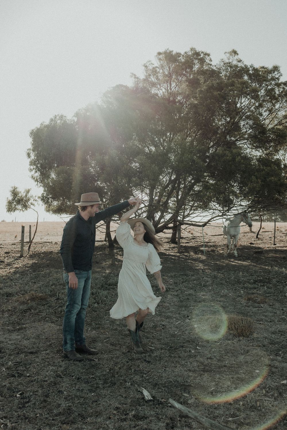 Man in cowboy hat spinning woman in white dress on a rural farm with sunflare through trees.