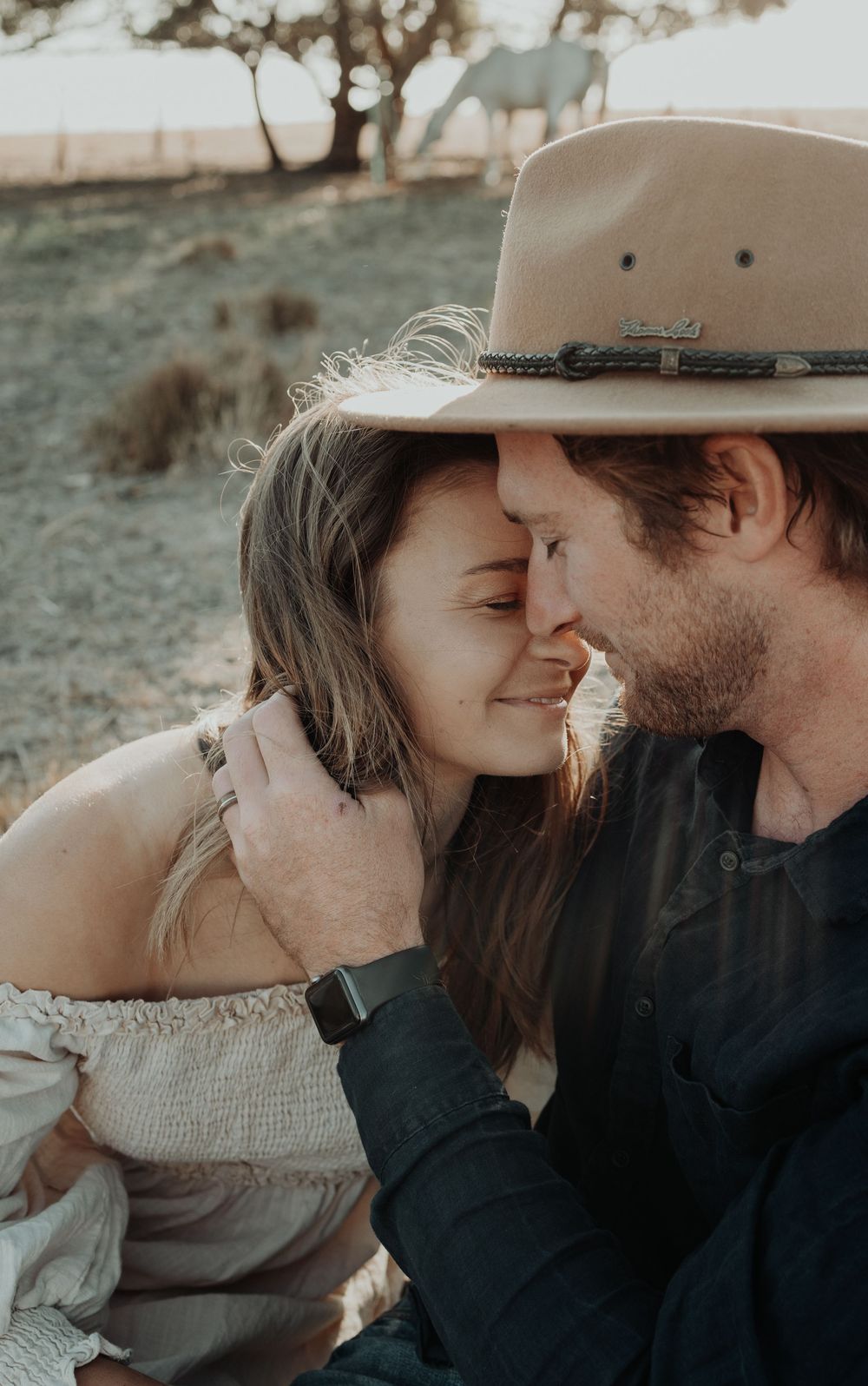 Intimate close-up of couple in cowboy hats sharing a tender moment with a horse in background.
