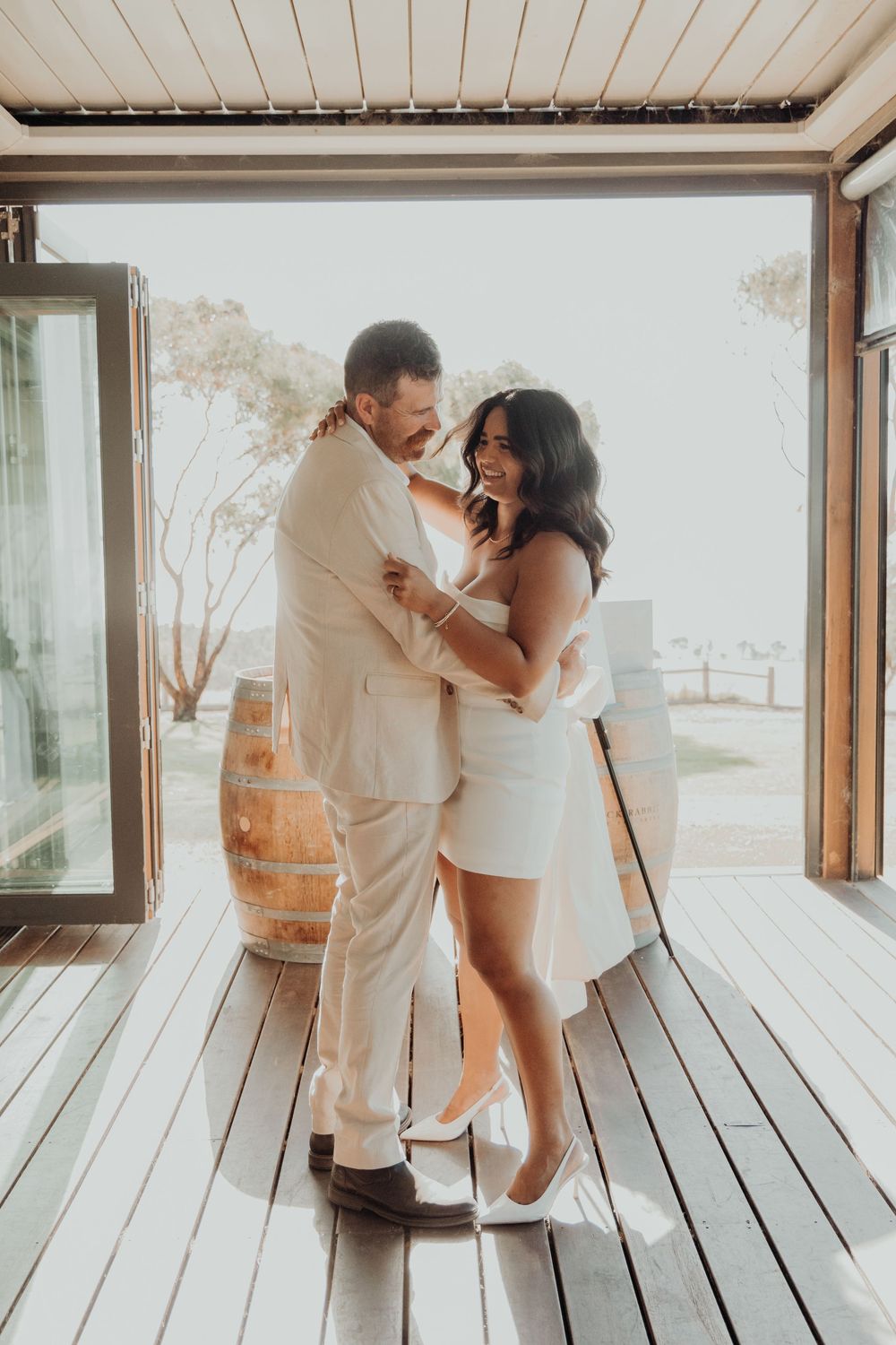 Bride and groom laughing and embracing on a wooden deck with warm backlit glow.