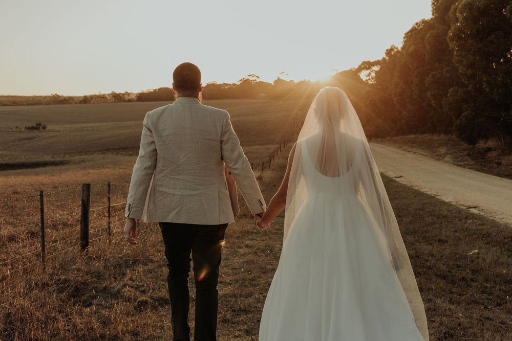 Bride and groom walk hand in hand along a rural path at golden sunset, bride wearing a long veil.