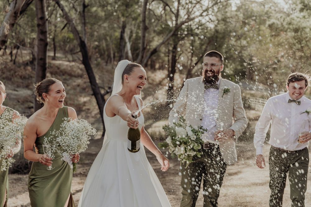 Joyful bride popping champagne bottle with bridesmaids in sage green dresses and groom in bush setting.