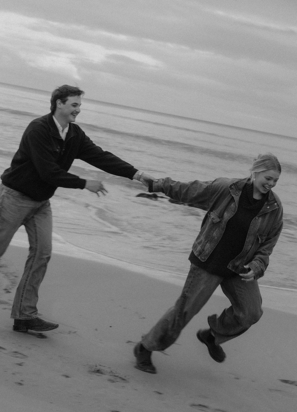 Black and white photo of a joyful couple holding hands and running playfully on a sandy beach with ocean waves behind them.