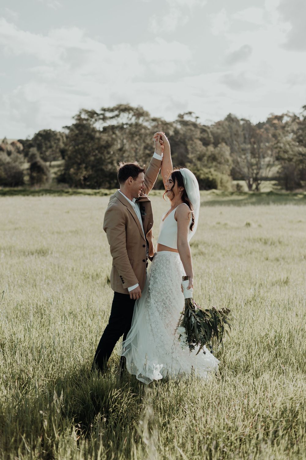 Groom spinning bride in a sunlit green field, both smiling joyfully during wedding portraits.