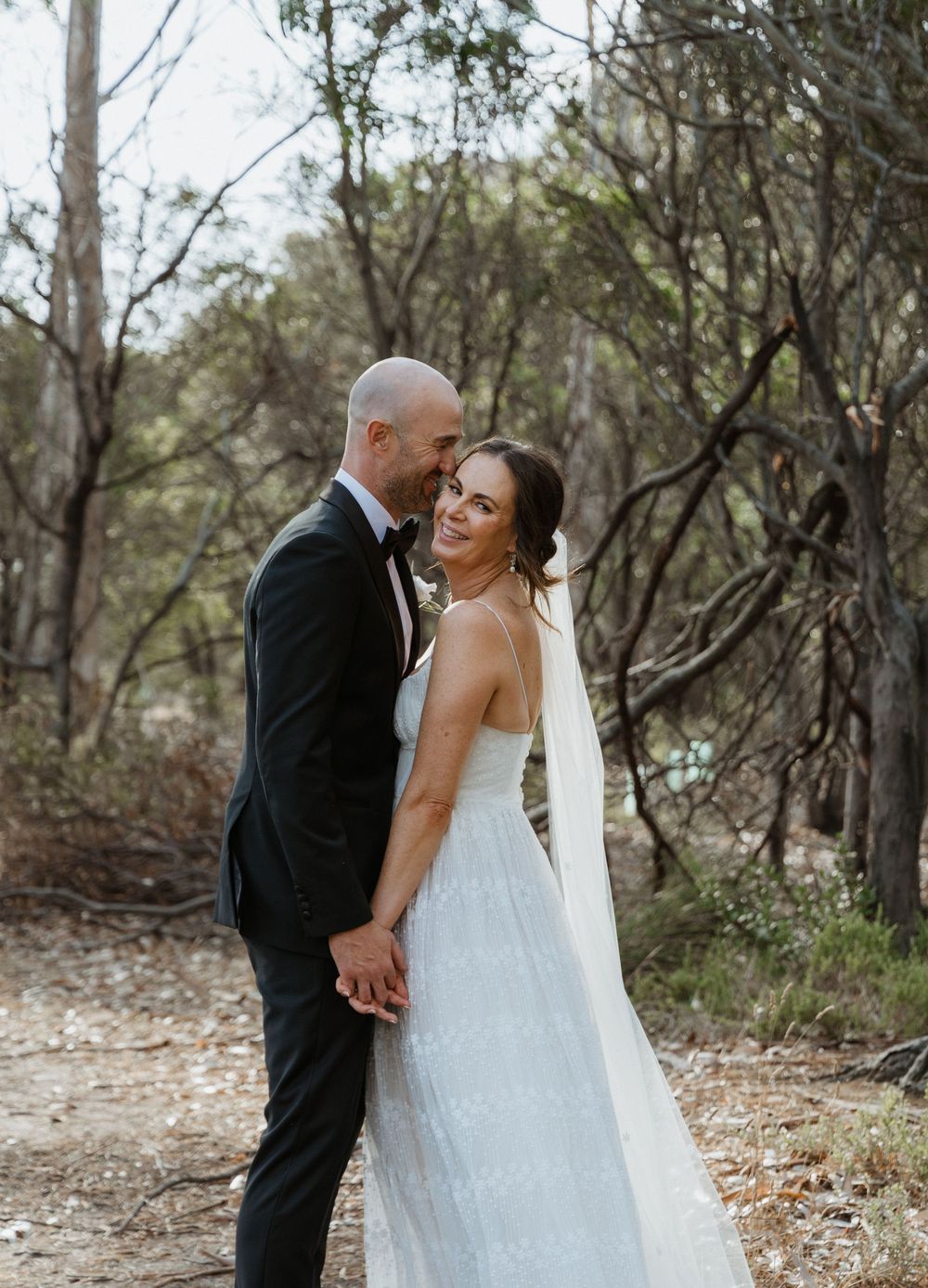 Groom kisses bride on the cheek as she smiles at camera in a lace wedding dress among bushland trees.