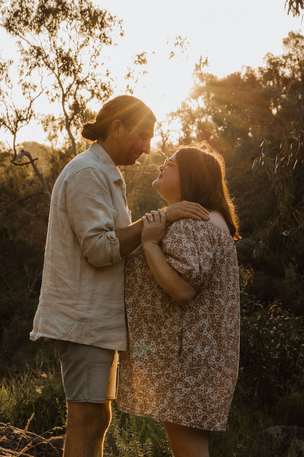 A couple shares an intimate moment face-to-face in golden sunset light surrounded by Australian bush trees.