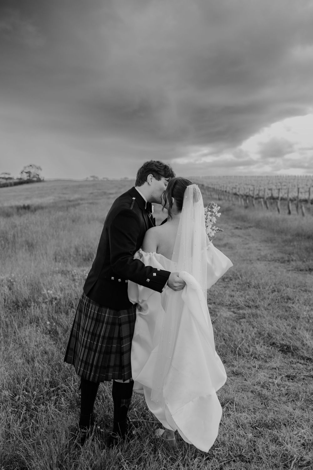 Bride and groom sharing romantic kiss in field under moody storm clouds, groom in tartan kilt.