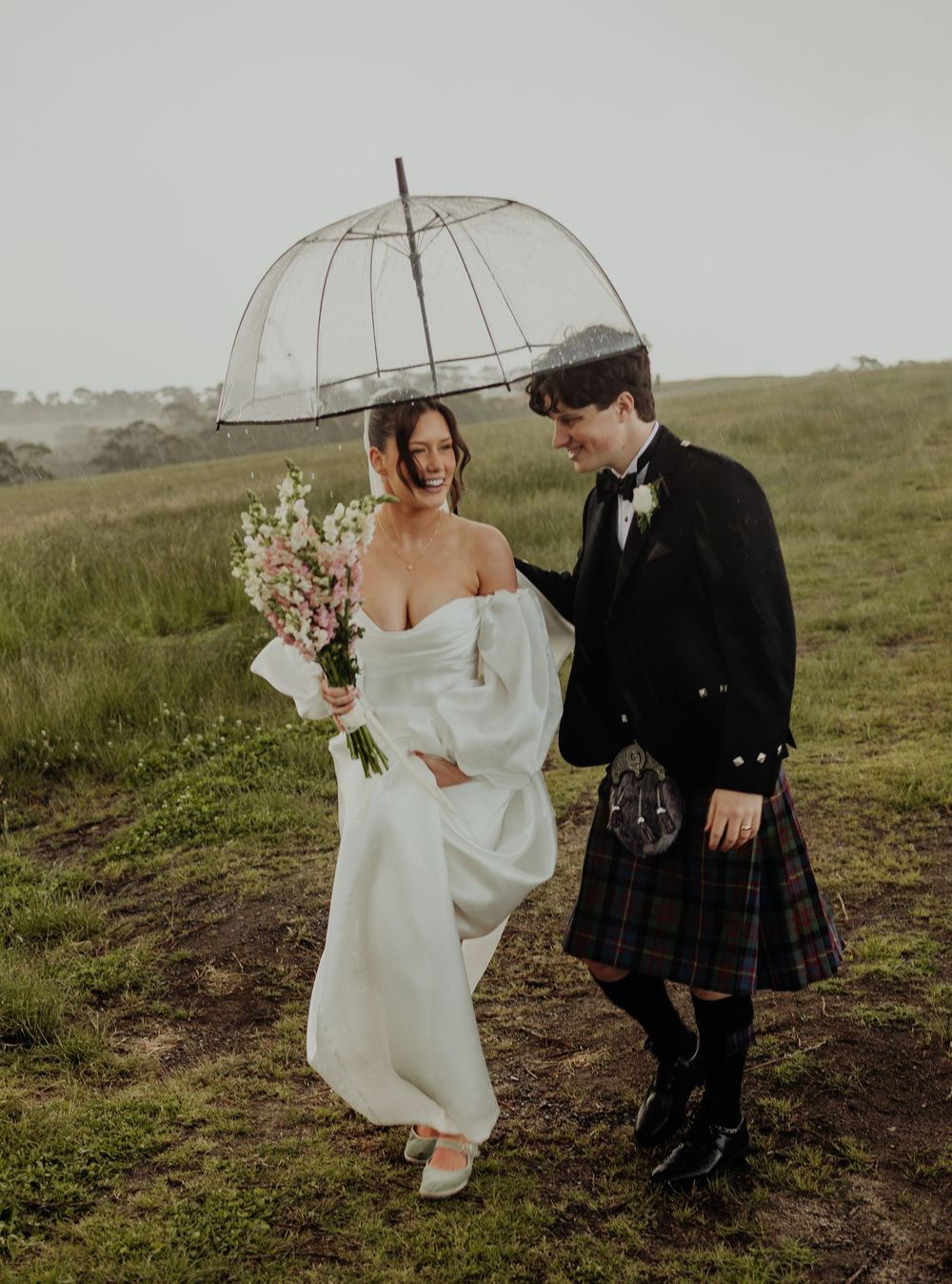 Laughing bride and kilt-wearing groom share a clear dome umbrella while walking through a rainy green field.