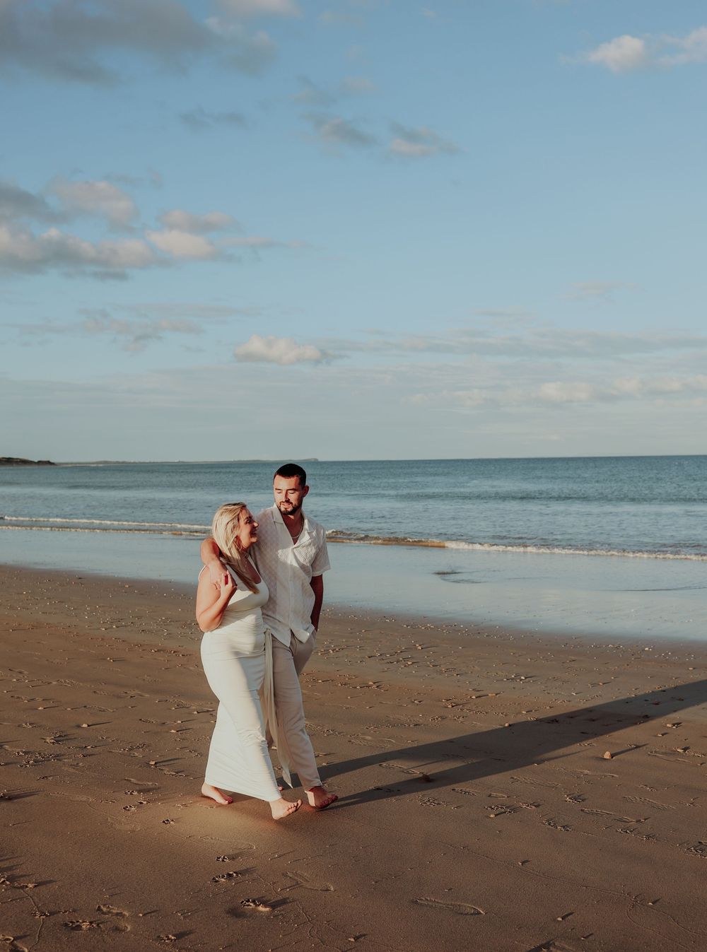 Engaged couple walking barefoot on beach at golden hour sunset.
