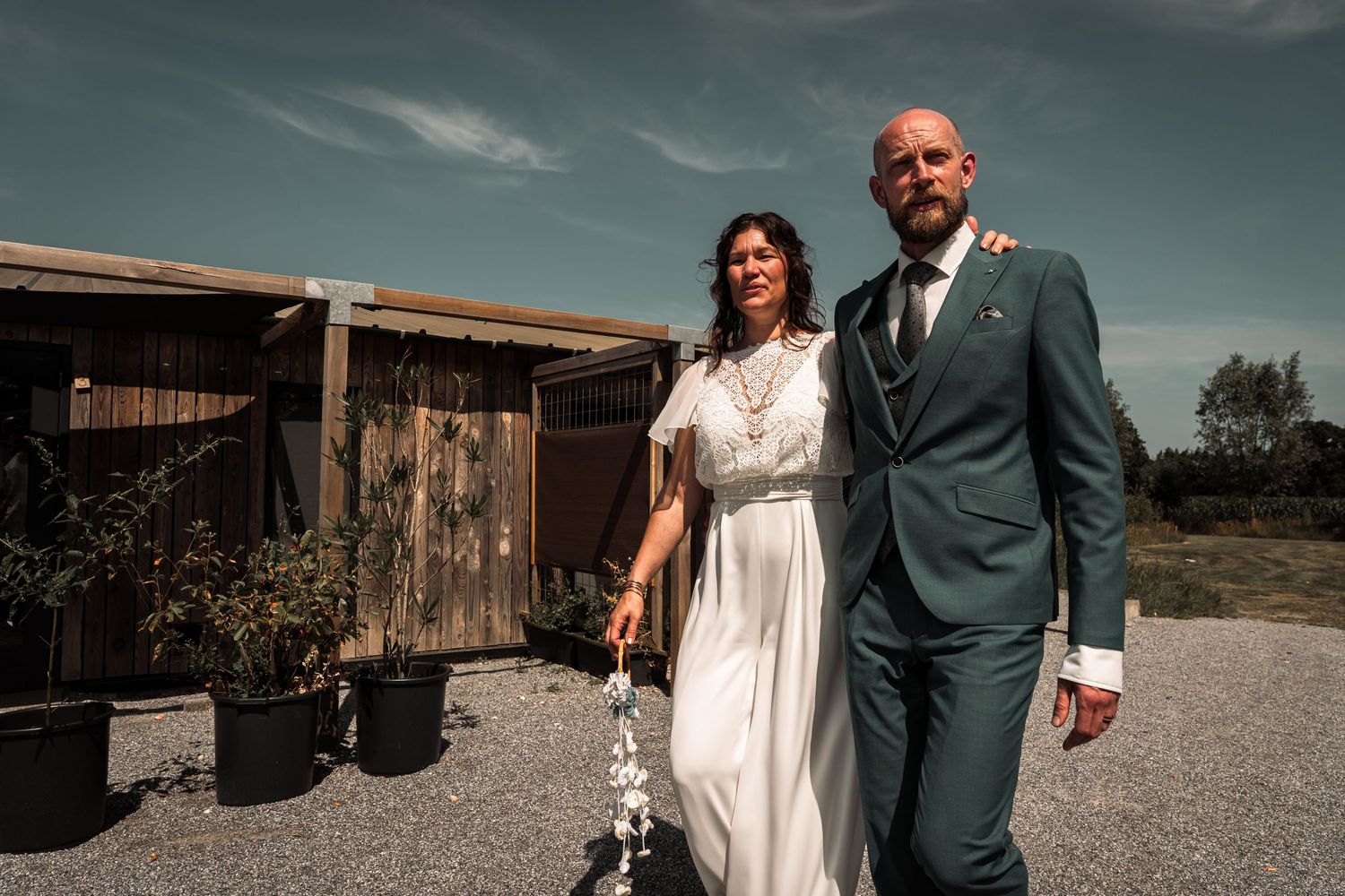 A couple in wedding attire stands together outdoors near a rustic wooden barn on a cloudy day.