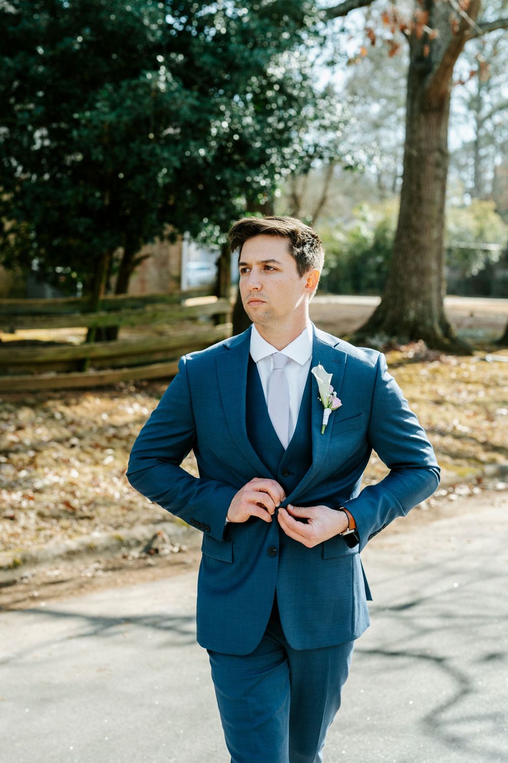 Groom in navy blue suit and white tie poses in an outdoor setting with autumn leaves on the ground.