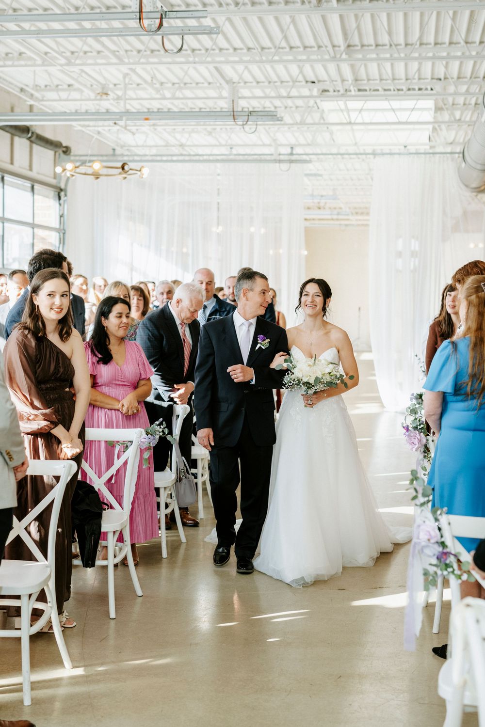 Wedding guests in formal attire gather in a bright indoor venue with white chairs and decorative elements.
