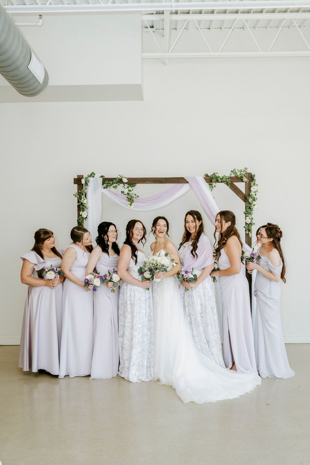 A bridal party in light lavender dresses poses together in front of a simple floral arch in a bright indoor space.