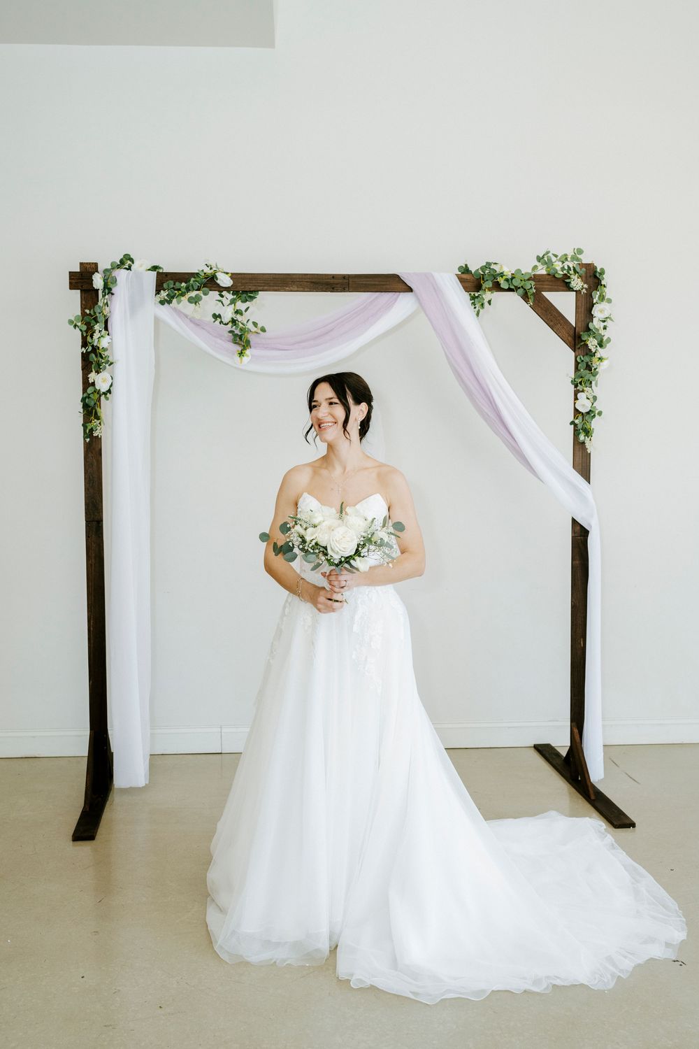 Elegant wedding setup with wooden arch decorated with greenery and flowing white fabric against light backdrop.