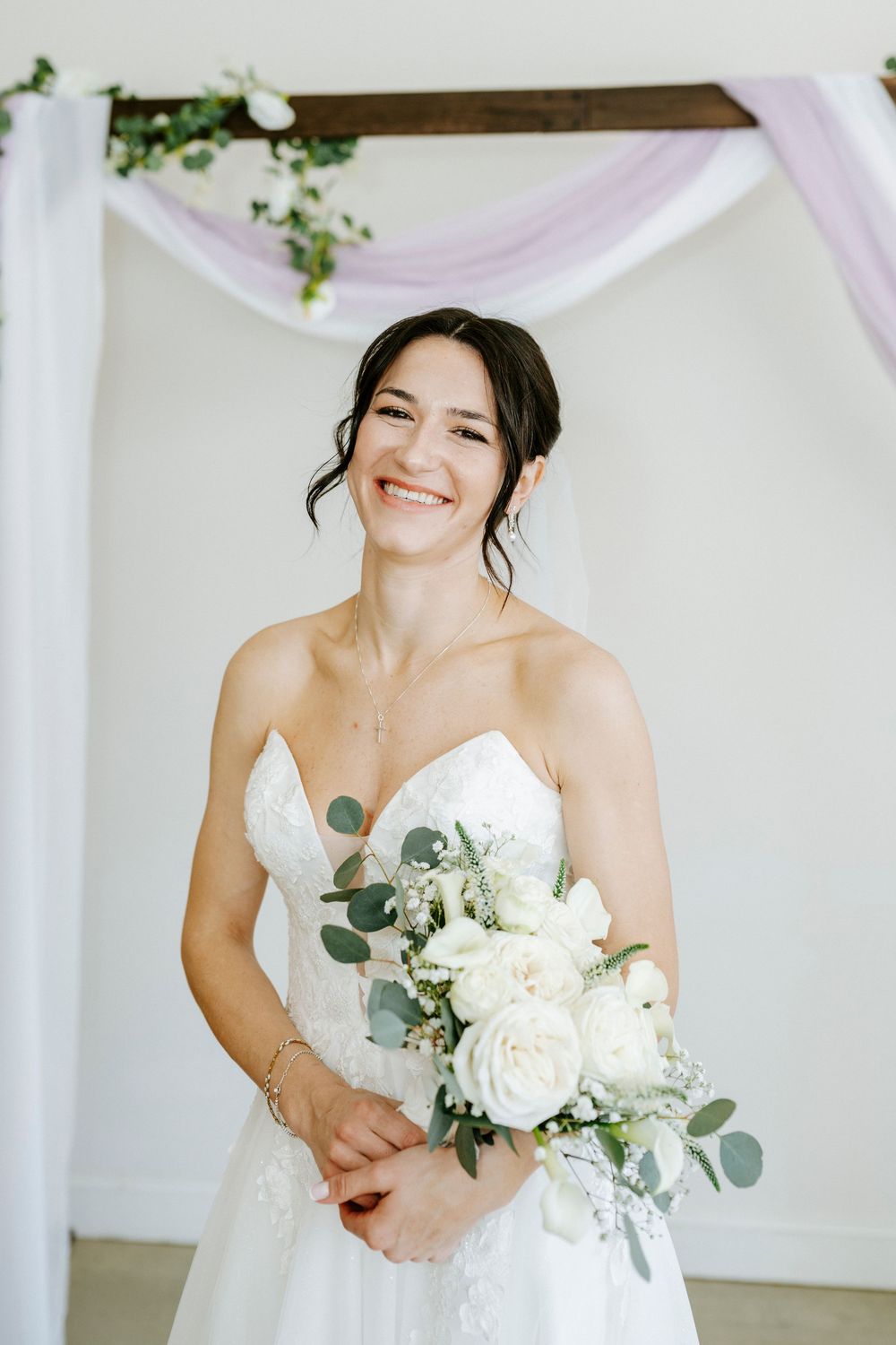 A bride in a strapless white wedding dress holds a beautiful bouquet of white roses and eucalyptus against a draped backdrop.