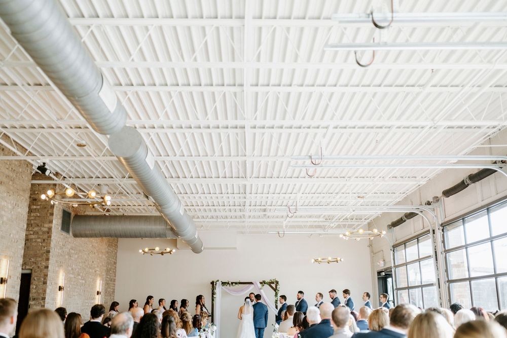Indoor wedding ceremony in an industrial-modern venue with white ceiling, exposed ductwork and large windows.