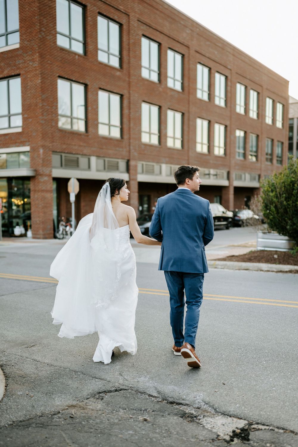 Bride in flowing white dress and groom in navy suit walk away from brick building on paved road.
